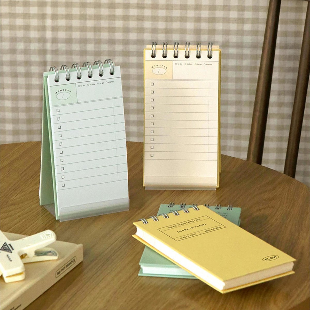 Two spiral-bound notepad books and a matching yellow hardcover notebook arranged on a wooden table, with a checkered curtain and wooden chair visible in the background.