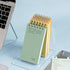 A stack of spiral-bound notepads in pale green and yellow stands on a clear acrylic desk pad on a light blue surface, with part of a laptop and a clipboard visible nearby.