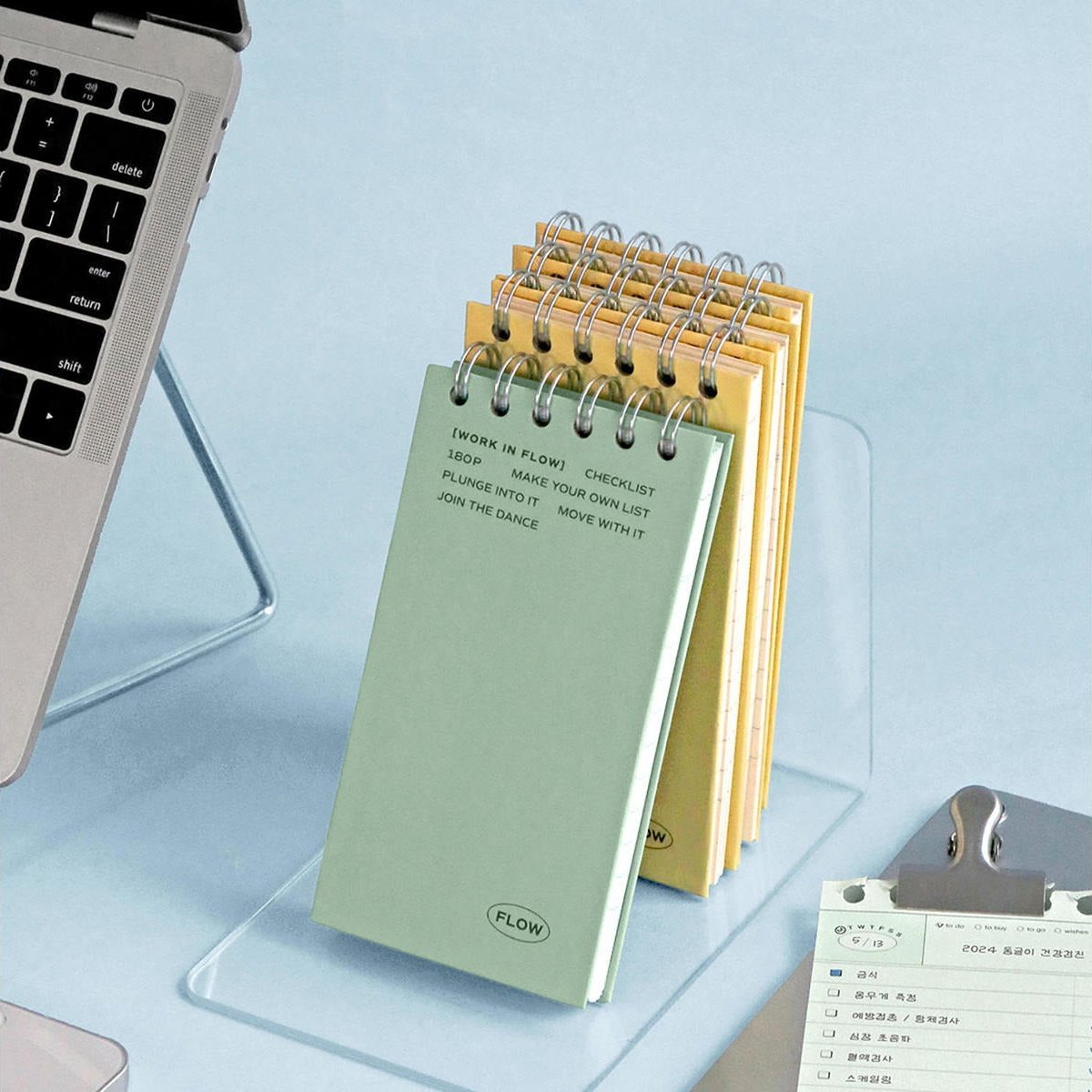 A stack of spiral-bound notepads in pale green and yellow stands on a clear acrylic desk pad on a light blue surface, with part of a laptop and a clipboard visible nearby.
