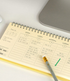 A spiral-bound planner page showing a weekly schedule layout in English and Korean, photographed on a light desk with part of a laptop visible and a gray pen resting on the page.