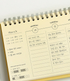 A spiral-bound planner page shown close up, with cream paper, silver metal spiral binding, and printed sections for Monday and Tuesday with handwritten notes in Korean.