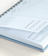 A close-up of a spiral-bound planner or notebook showing light blue lined pages with printed daily planning boxes and a calendar section, resting on a light neutral surface.