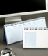 Two spiral-bound desk planners with light blue covers and white pages are displayed on a gray desk in front of a computer monitor. One planner is open to show a weekly schedule page, and the other is open to show a grid checklist page.