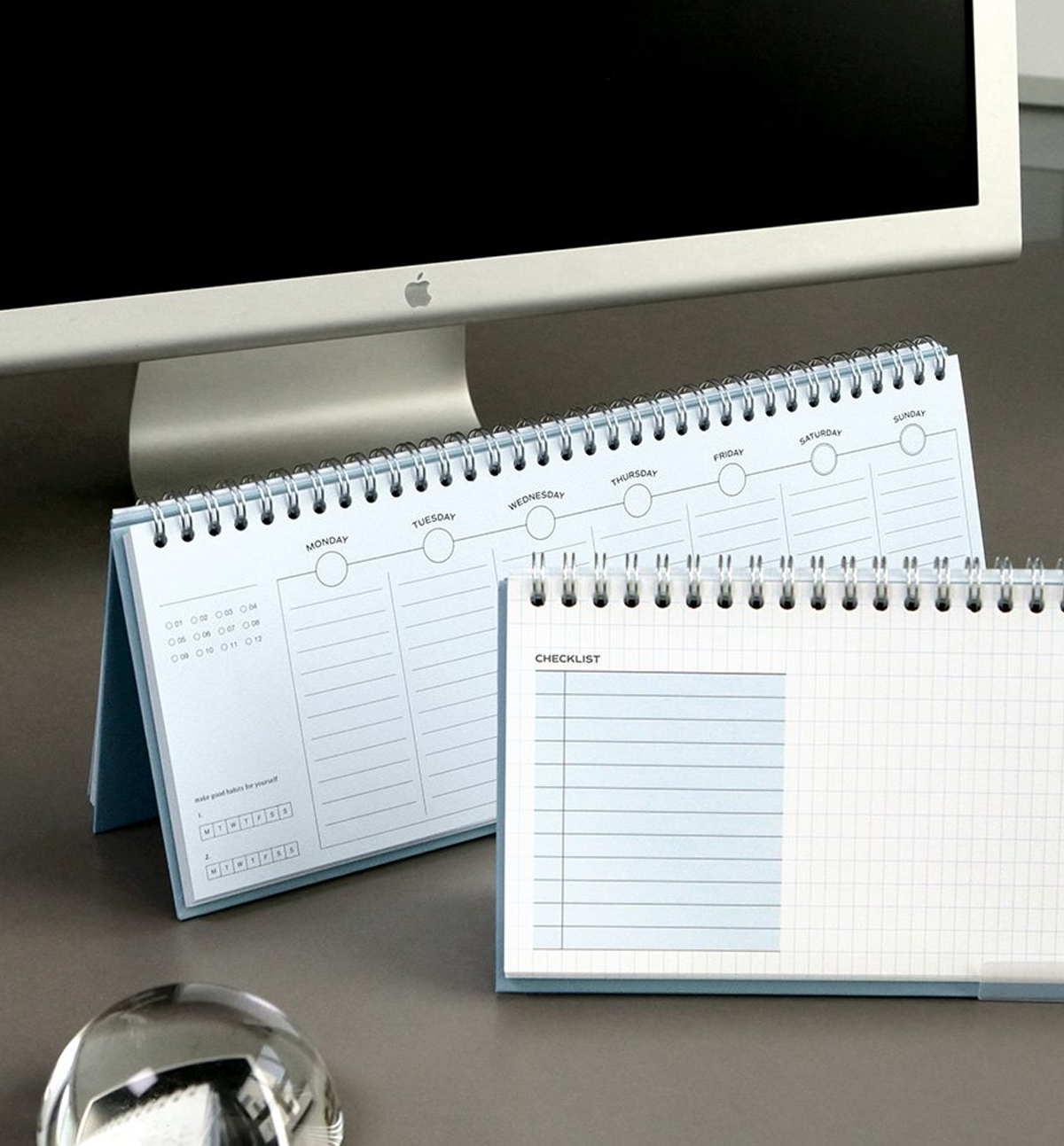 Two spiral-bound desk planners with light blue covers and white pages are displayed on a gray desk in front of a computer monitor. One planner is open to show a weekly schedule page, and the other is open to show a grid checklist page.