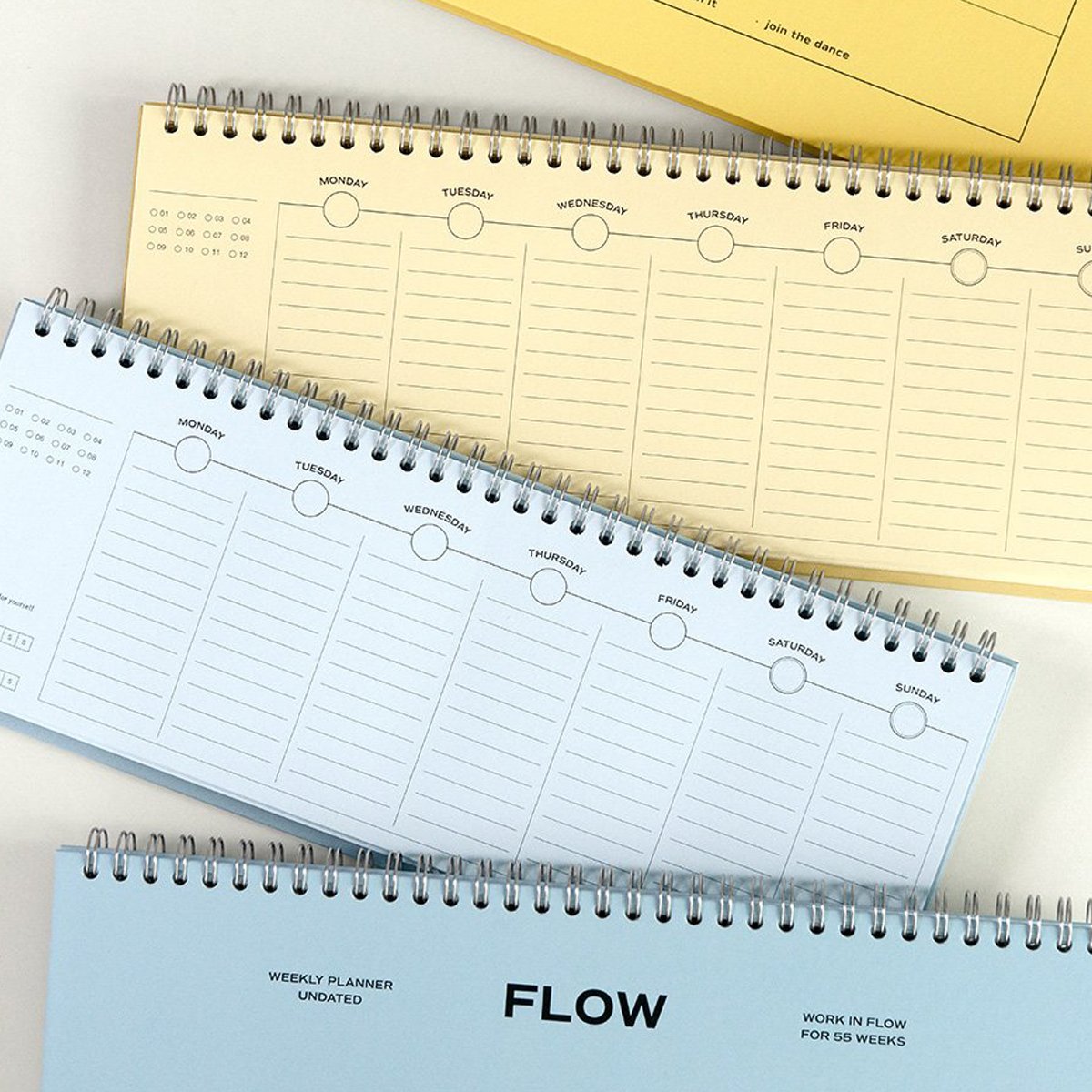 Three spiral-bound weekly planner notebooks in pastel blue, cream, and yellow, arranged diagonally on a white surface. The bottom notebook features the brand name 'FLOW' printed prominently on its cover.