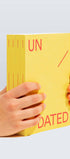 Yellow book titled 'UNDATED' held by a hand on a light gray background